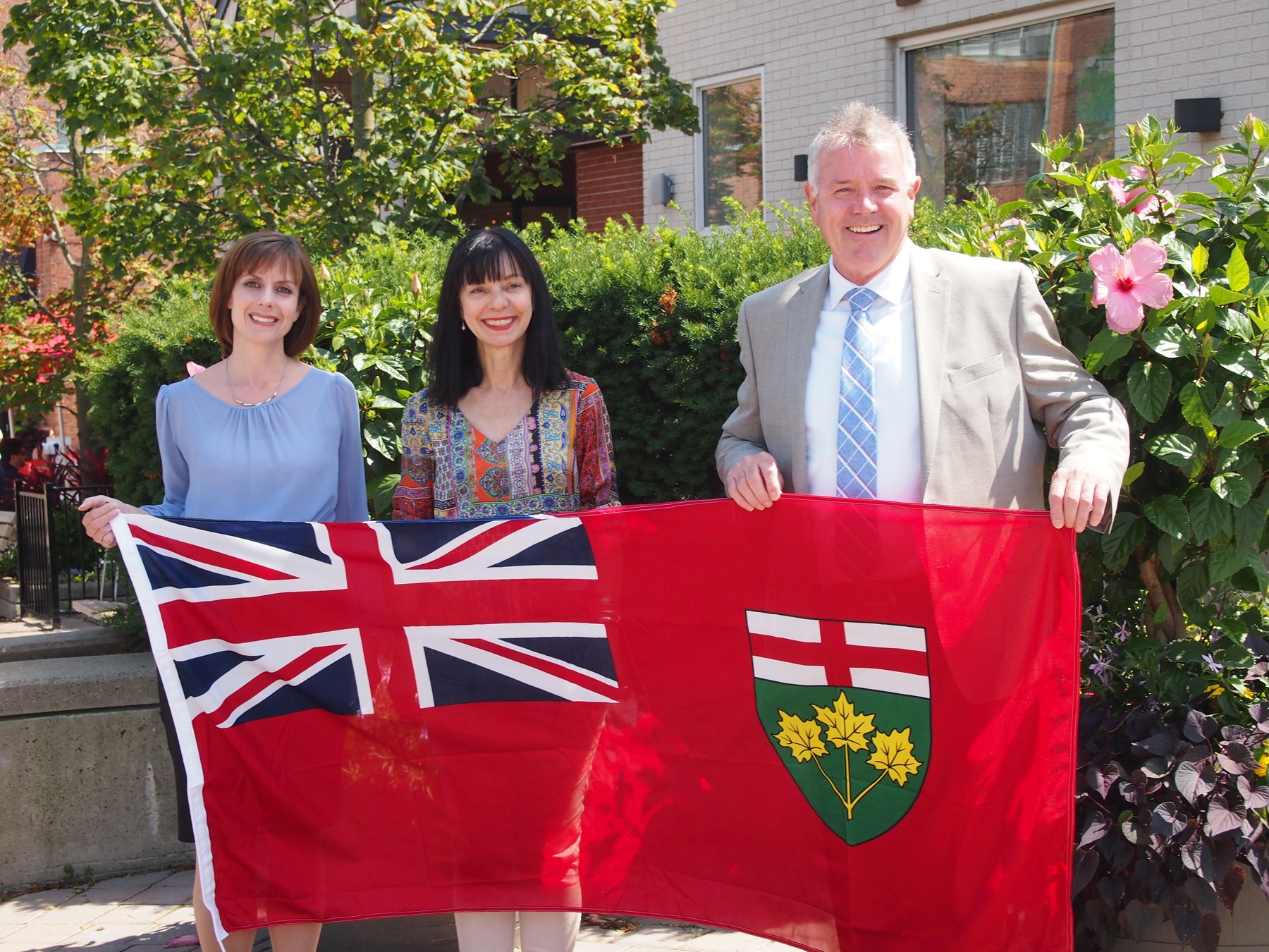 Two women and a man hold up the Ontario Provincial Flag | MPP Oakville Ontario