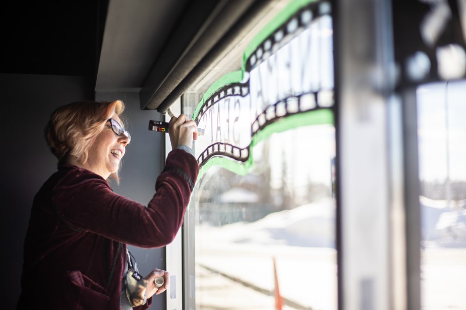 Okotoks artist Robin Thibodeau paints a mural in the front windows of Okotoks Cinemas on Feb. 27 as a way of pitching in to help advertise their crowdfunding campaign for new theatre seating.