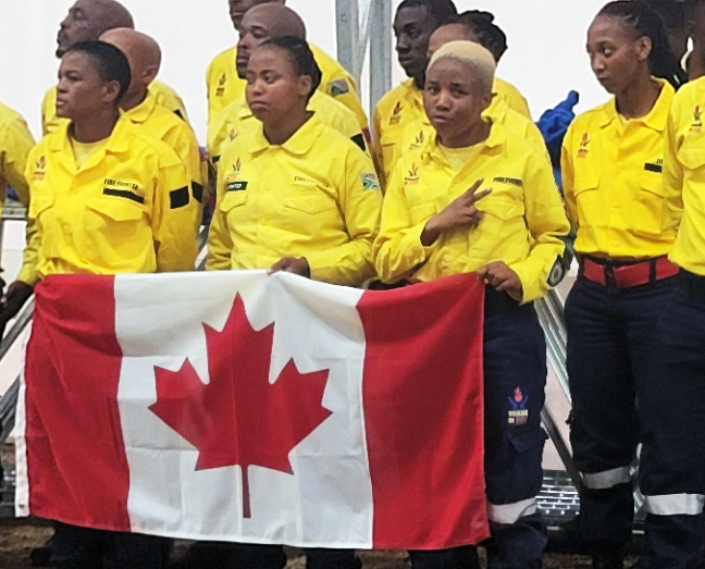 Three female firefighters from South Africa hold the Canadian flag while they sing their country's national anthem during a ceremony Thursday at their fire camp near Prince George Airport.