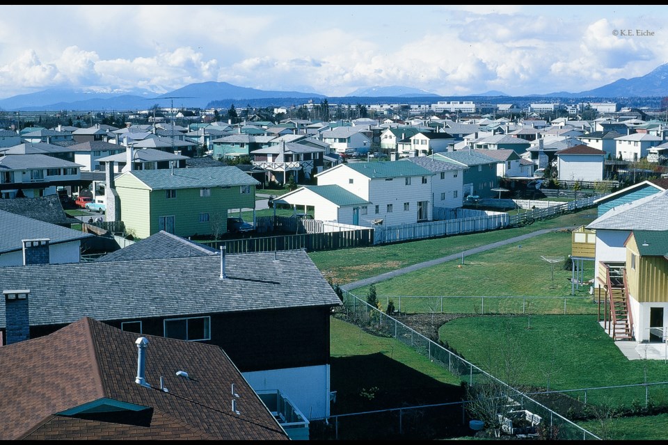 View towards north-west. The footpath through the grassy area seen at right leads from the end of Tranquille Place to the Samuel Brighouse school grounds. Kalamalka Crescent is the street visible at centre left. Beyond the houses lies Vancouver International Airport, with the main terminal (inaugurated 1968) and its air traffic control tower visible towards the centre in the top third of the photo. Some of the buildings further to the right are on the site of the old airport, now called the South Terminal. 