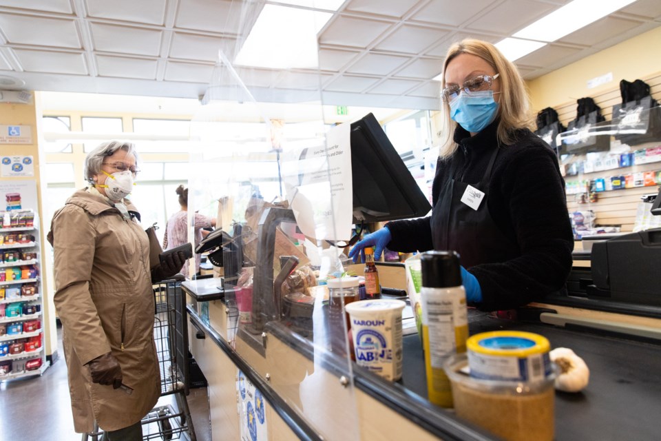 Karen Himmaugh rings up Barbara Bunker's groceries at Piazza's Fine Foods in Palo Alto on April 9, 2020. The two are separated by a large piece of plexiglass. 