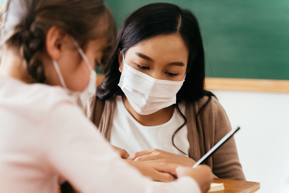 Close-up of Asian female teacher wearing a face mask in school building tutoring a primary student girl. Elementary pupil is writing and learning in classroom. 