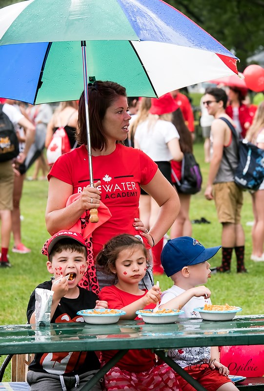 The first of several afternoon rain showers arrived just as the parade was coming to an end.  Here, Sarnia's Cori Savo provides a sheltering umbrella for, from left, Andrea Savo, 4, Avaya Savo, 6 and Brennen Penner, 4, eating Italian pasta. Glenn Ogilvie 