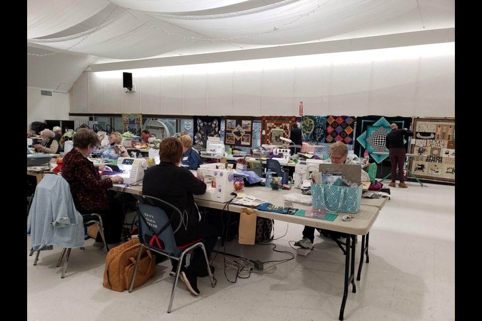 Quilting enthusiasts gather to in an event known as Quilt 'Til You Wilt, held in Unity, with finished products displayed behind work tables.