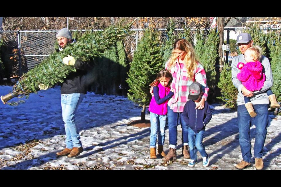 Steve Burris of the Weyburn Young Fellows carried a seven-foot tree over to the shack for payment, as the Bakken family made their selection on Saturday. Amanda Bakken is with daughter Embry and son Bryson, and Marshall is carrying his youngest daughter, Jemma.