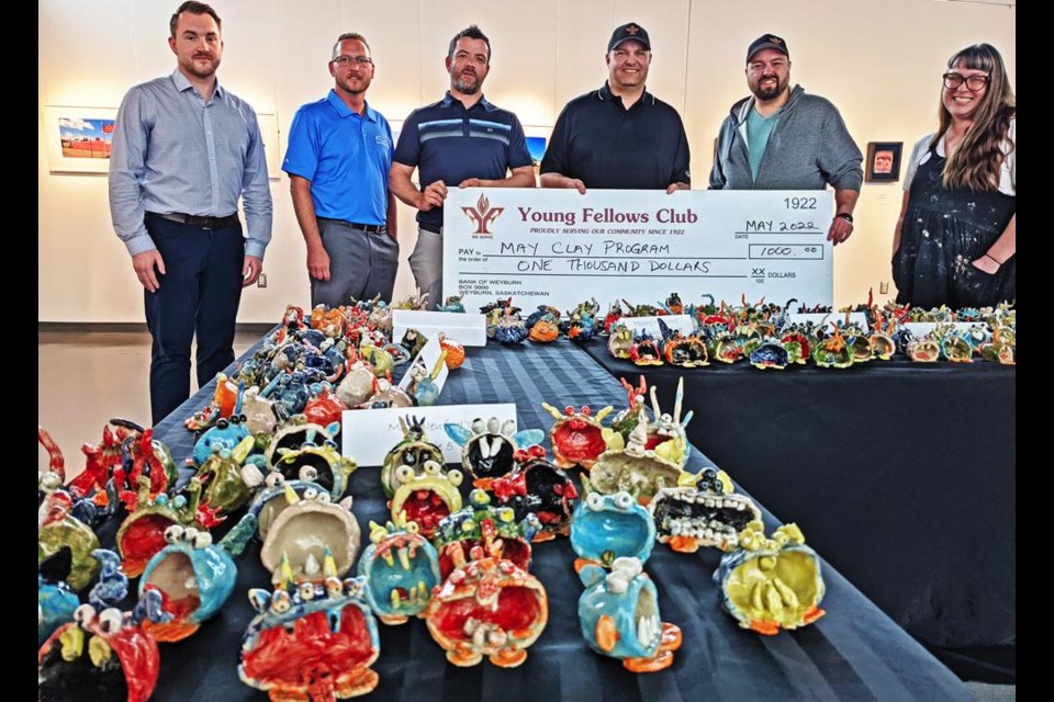 Members of the Young Fellows gathered to present a $1,000 donation to Regan Lanning for the May Clay program with finished examples of the projects in the foreground in the Weyburn Art Gallery