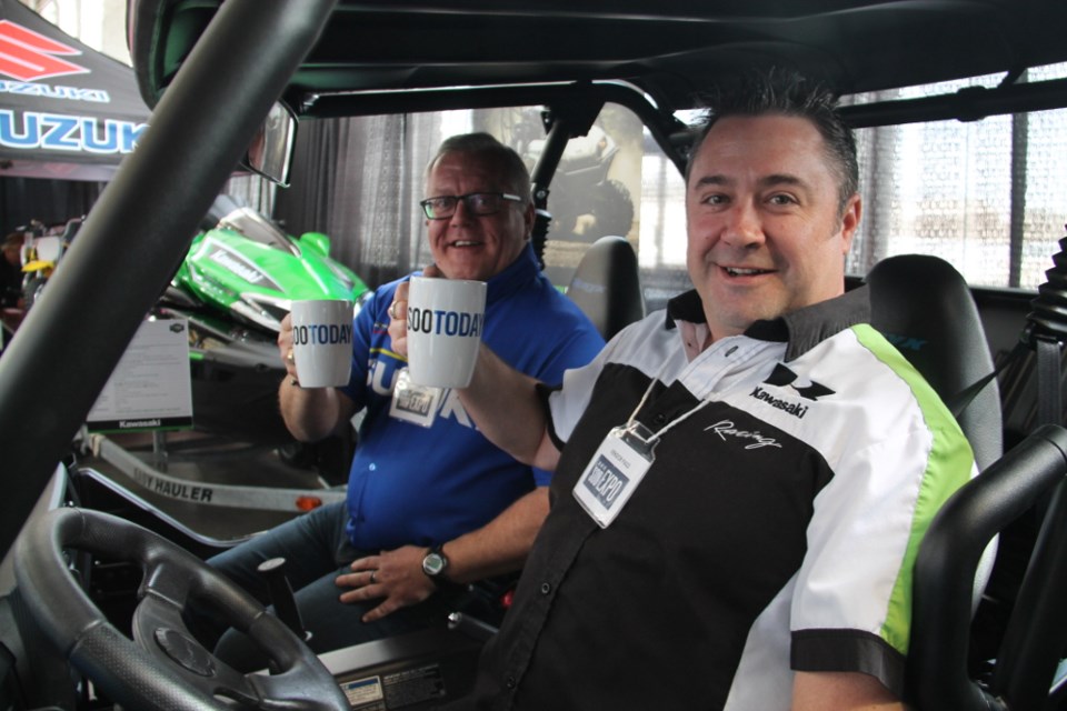 Greg Robinson, Bud Robinson Motorsports president/dealer principal (at the wheel of a Side by Side) and Scott Livingstone, Bud Robinson Motorsports sales consultant, with their complimentary SooToday coffee mugs at the Sault Ste. Marie Chamber of Commerce Big Sport and Outdoor Show held at The Machine Shop, March 10, 2018. Darren Taylor/SooToday