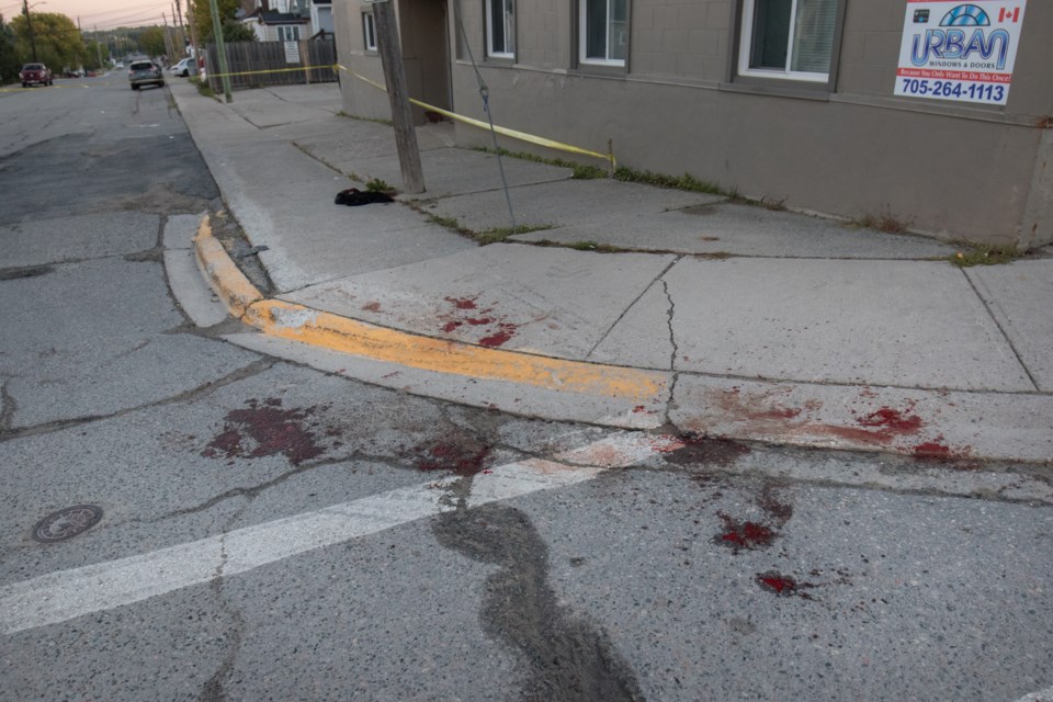 Blood and articles of clothing can be seen on the sidewalk and road behind police tape the intersection of Elm Street North and Algonquin Street East this morning. Jeff Klassen/TimminsToday