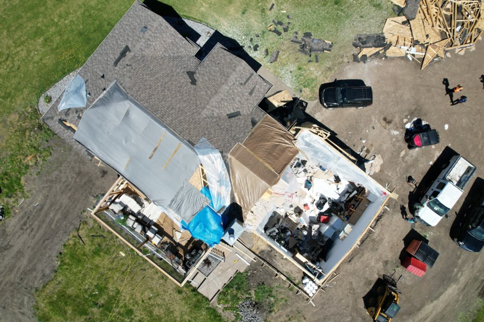 A close up aerial shot of one of several houses left without parts of its roof, debris on the ground, and clean up crews and equipment. 
