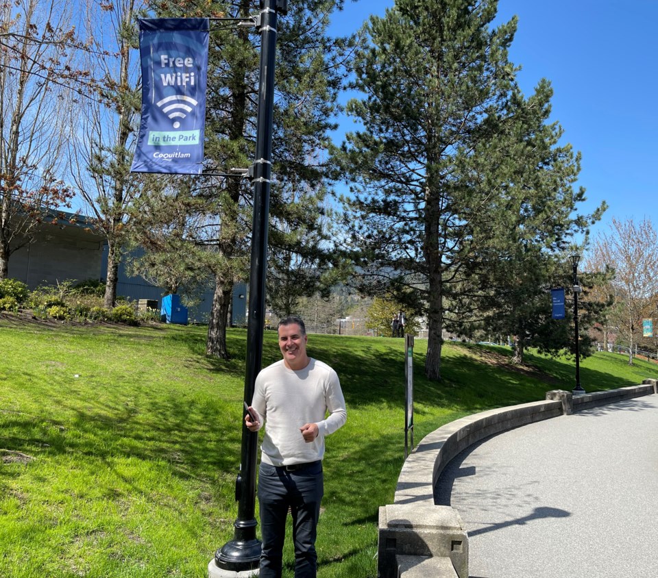 Danny Bandiera, City of Coquitlam’s Manager of Information and Communications Technology, stands in front of a blue Coquitlam-branded banner that says “Free Wi-Fi in the Park” while holding up a cellphone in his left hand in Town Centre Park.