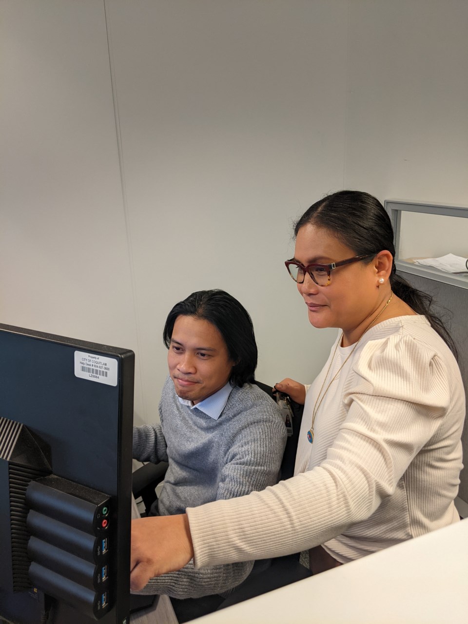 An image of Infrastructure Manager Darlene Soque in a white shirt standing beside a colleague in a gray sweater who is sitting behind a computer. Darlene’s left arm is raised to point to something that is on the screen in the image.