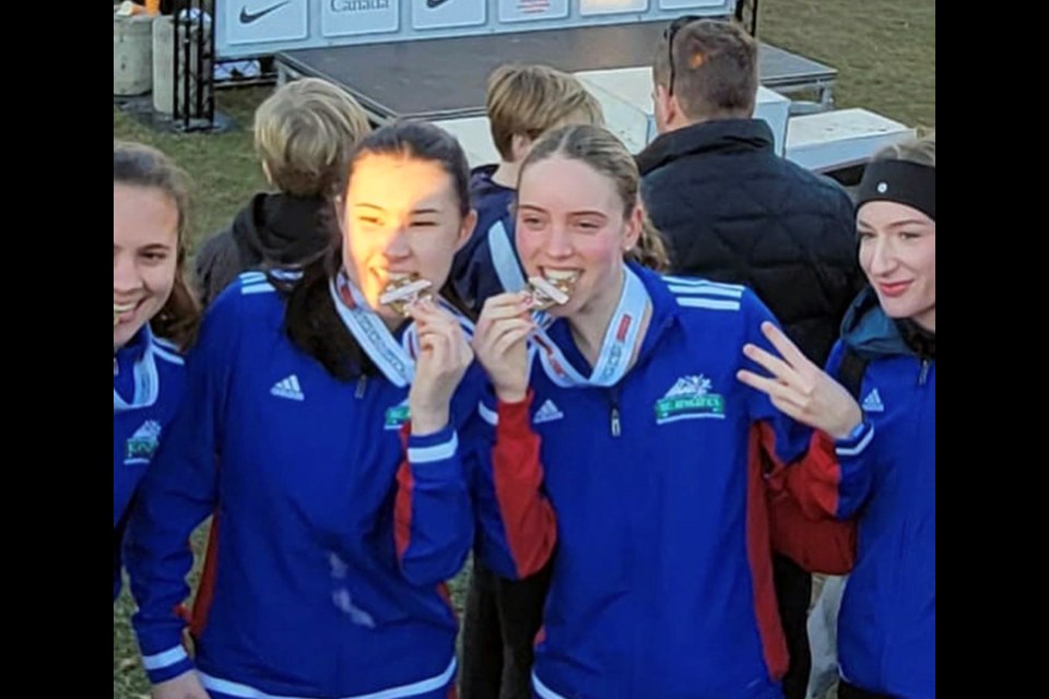 Coquitlam runners Ella Kalnins (left) and Ella Madsen bite into their gold medal with Team B.C. at the 2022 Canadian Cross Country Championships in Ottawa.