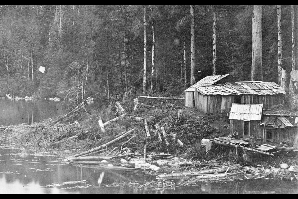 A Squamish home on the shores of Coal Harbour in 1868. A man sits on the front porch of one of the buildings, above the canoes. Reference code: AM54-S4-: St Pk N4