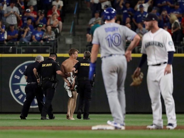  Toronto Blue Jays' Brandon Drury (10) stands on second base as a man who jumped out of the stands and ran naked in the Safeco Field outfield is taken away by sheriff's deputies during the ninth inning of the Blue Jays' baseball game against the Seattle Mariners, Saturday, Aug. 4, 2018, in Seattle. U.S. authorities say a Vancouver resident who ran naked onto a baseball field during a game in Seattle is set to appear in court today to face a criminal trespassing charge. THE CANADIAN PRESS/AP/Ted S. Warren